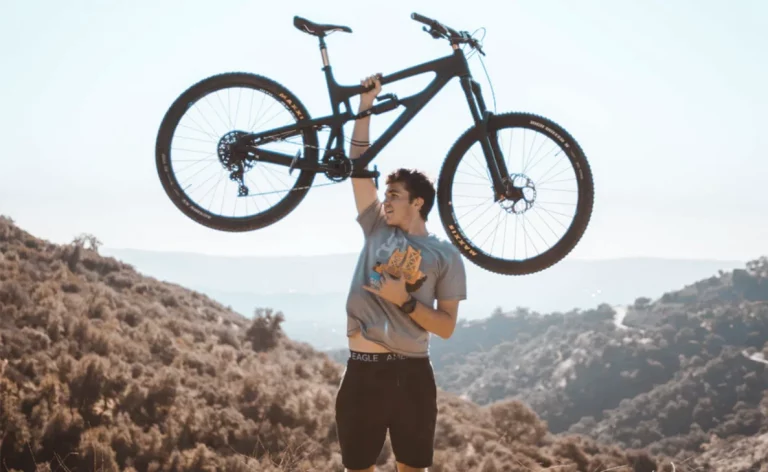 A man holding up a bicycle with one hand into the sky amongst a sandy background.