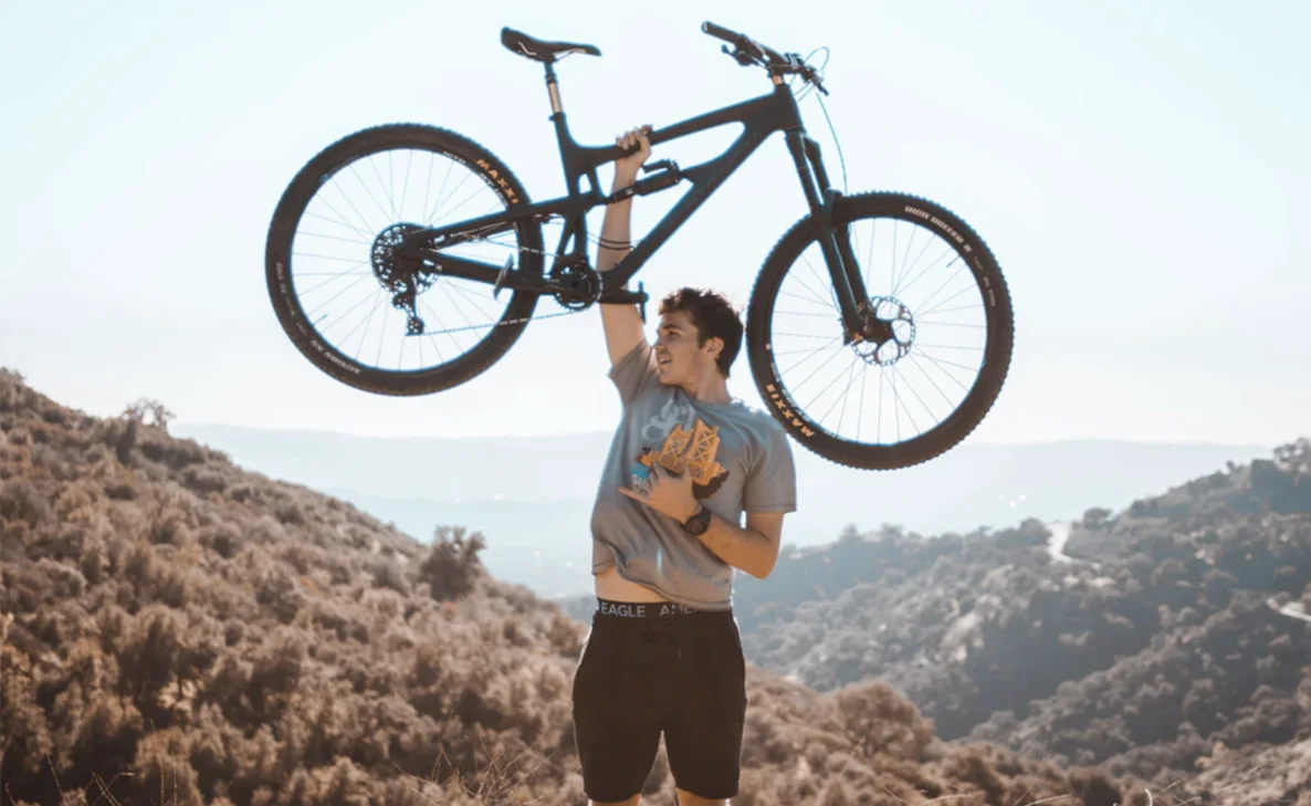 A man holding up a bicycle with one hand into the sky amongst a sandy background.