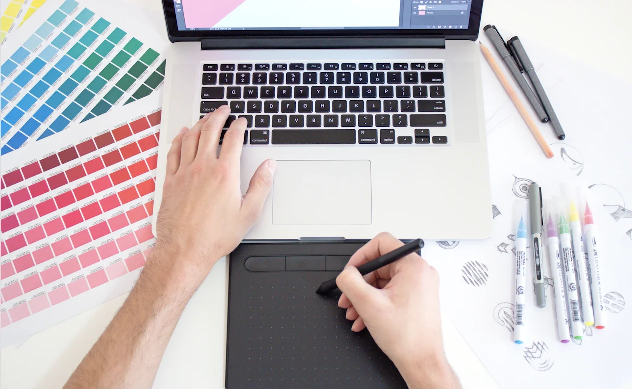 An image of a person using a drawing tablet with their laptop. Artistic tools are scattered around the table it is on.